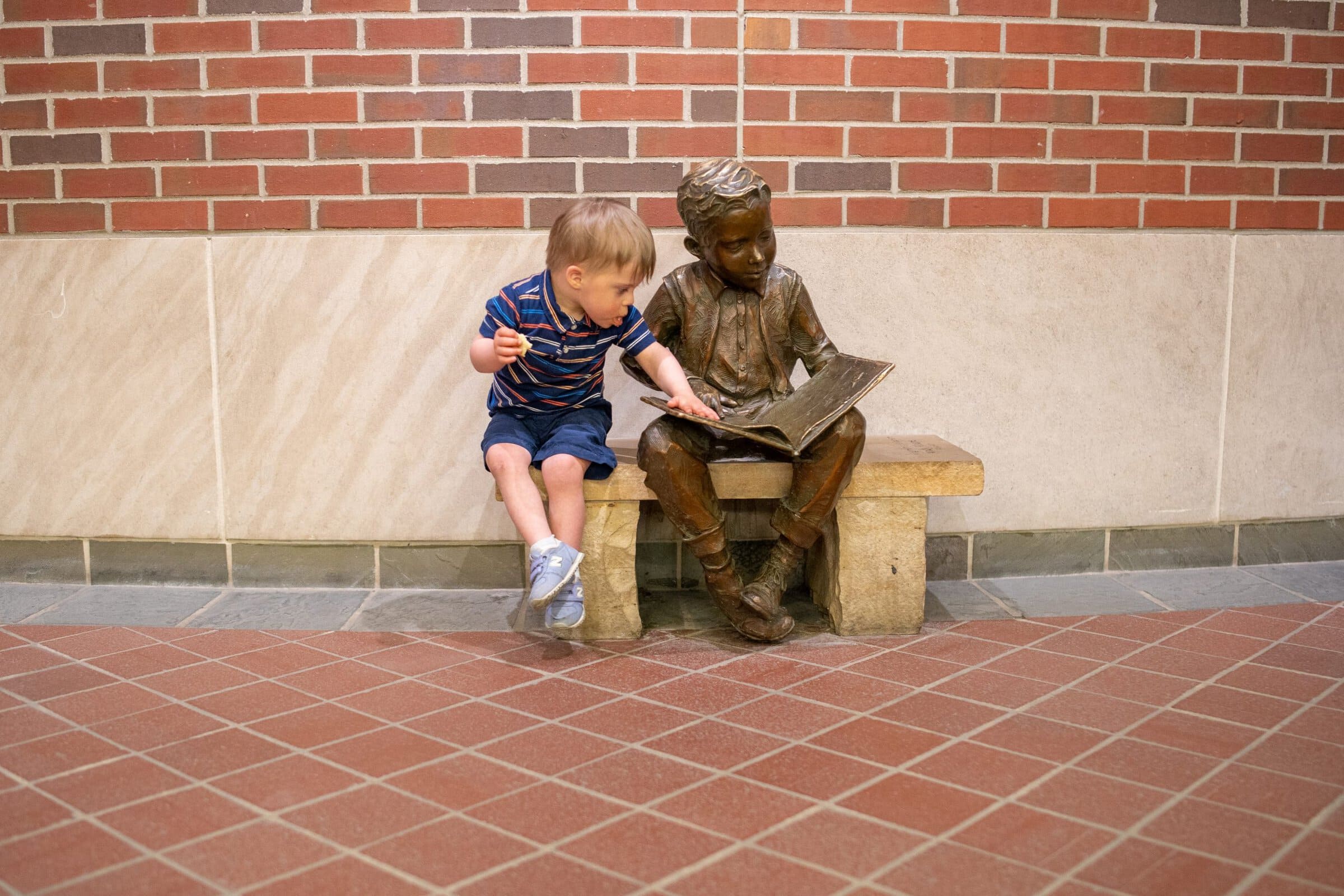 Young boy sitting next to the library statue of a boy reading on a bench.